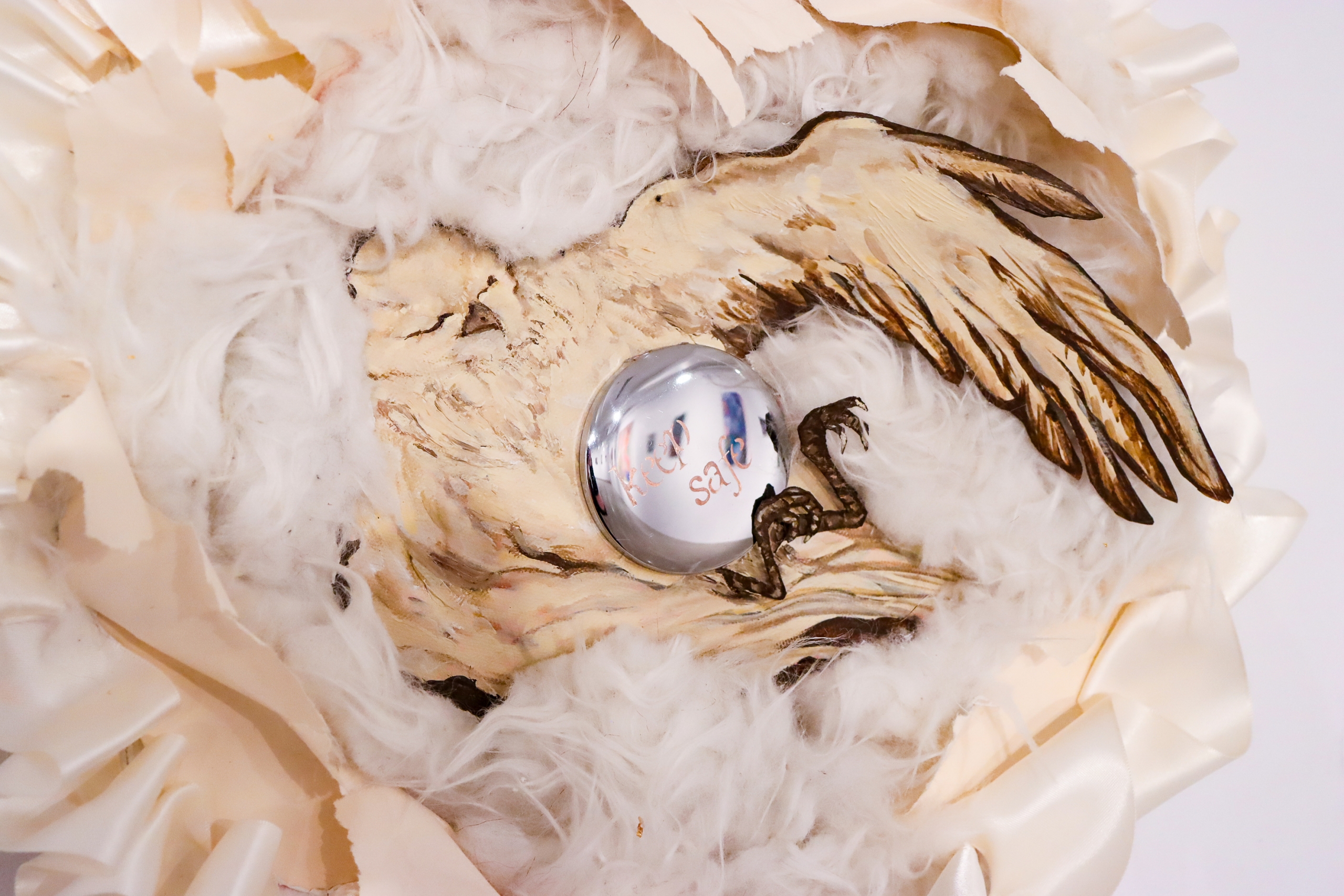 Close-up of a mixed-media sculpture: a pyrography bird cradling a mirrored silver sphere in soft feathers.