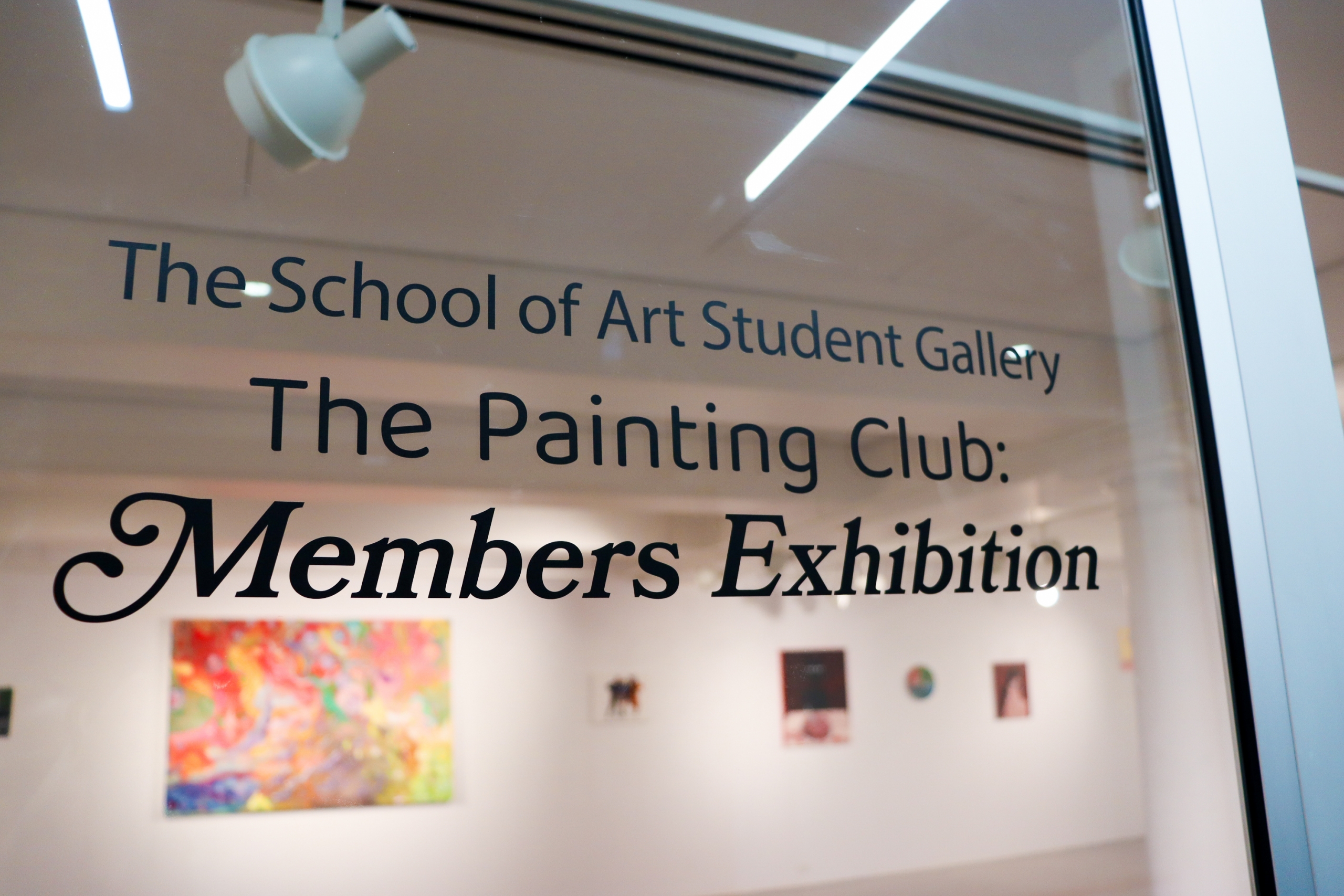 View through the gallery's glass entrance door showing the exhibition title vinyl lettering and colourful paintings on the walls beyond.