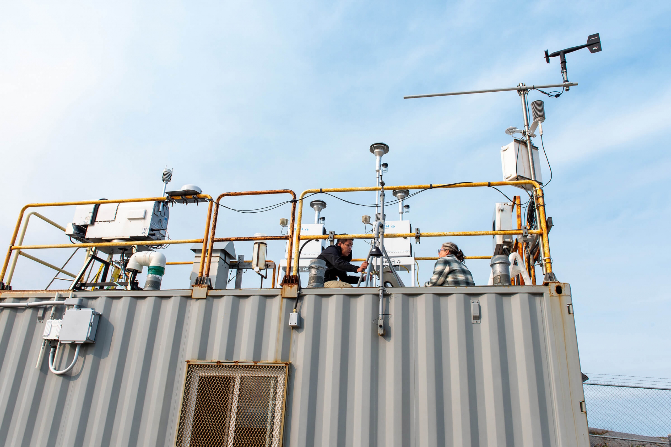 A man and a woman sit on the roof of a trailer adjusting scienctific equipment