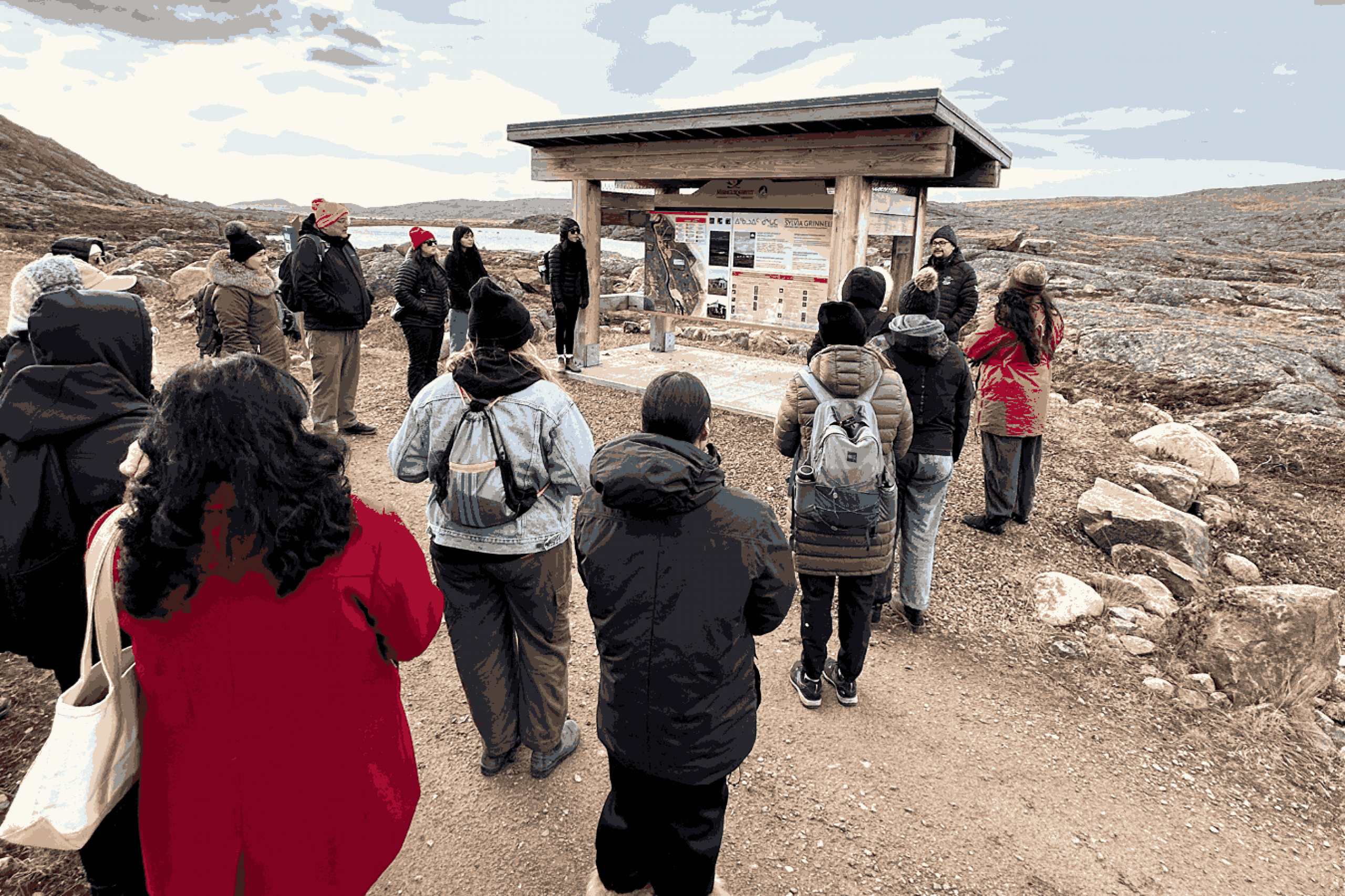 A group of people stand outdoors listening to a guide beside an informational sign near rocky terrain and water.